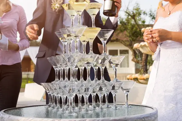 A groom fills a pyramid of champagne glasses at an outdoor wedding ceremony